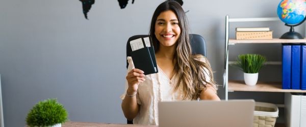 I am here to help you with your booking and travel questions. Female sales agent holding plane tickets while smiling and making eye contact at the travel agency